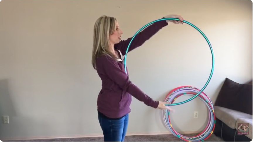 Michele McCoy demonstrating the continuous K-roll hoop trick with a teal hoop indoors.